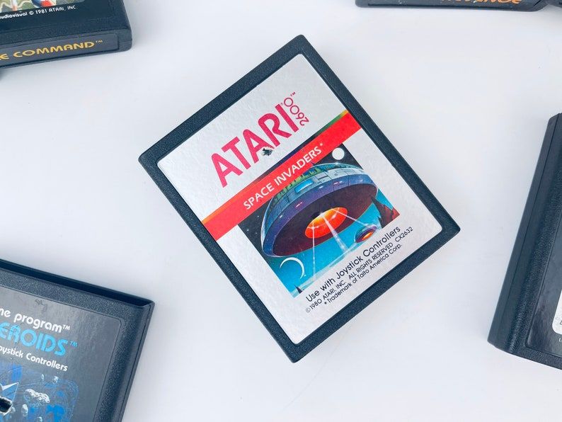 May include: A black Atari 2600 game cartridge for Space Invaders. The cartridge has a red and white label with the game title and instructions to use with joystick controllers. The label also includes copyright information for Atari, Inc. 1980.