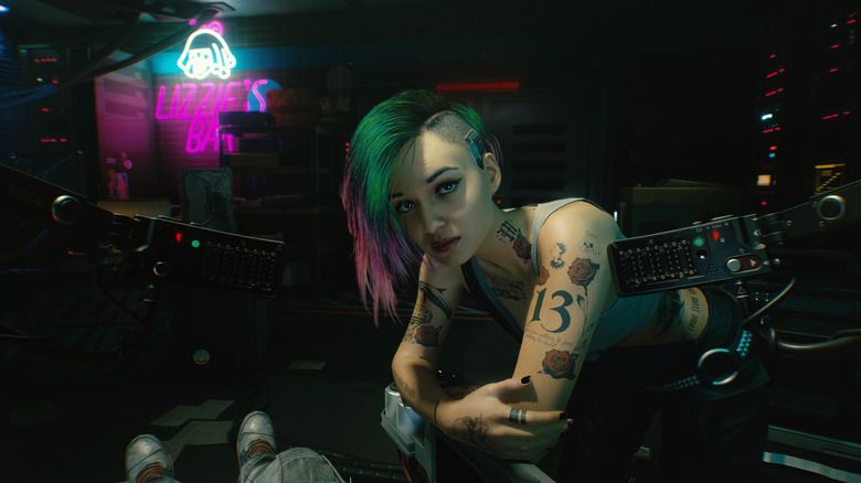 Screenshot from the video game Cyberpunk 2077 of the NPC Judy. A tattooed woman in a tank top leans on the arm of a futuristic medical chair in a dark room, with a neon sign in the upper left corner..